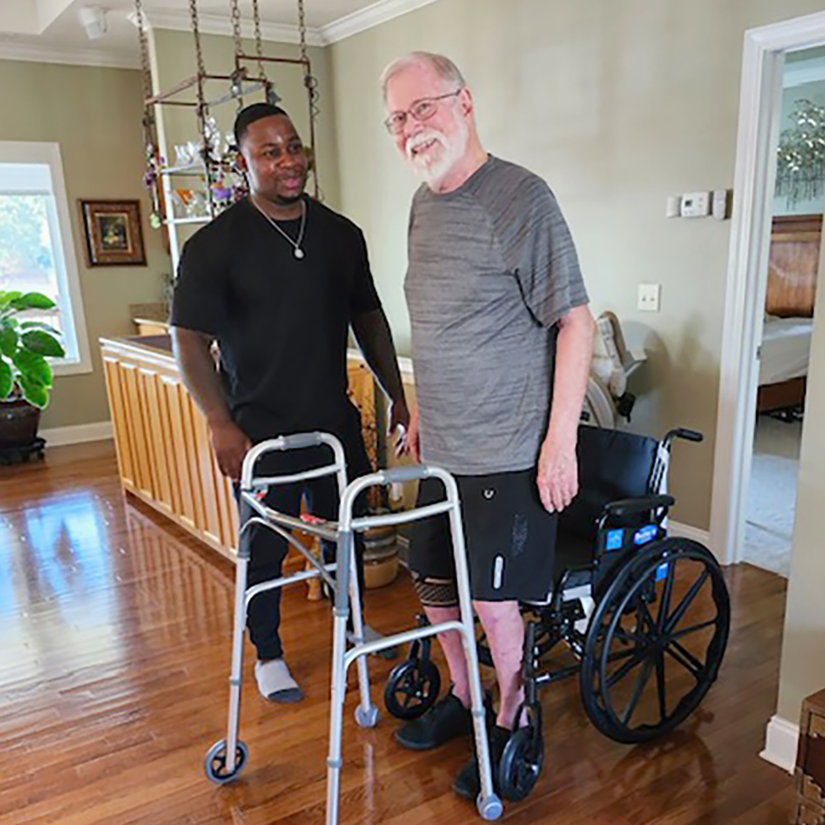 Two men are indoors; one is standing beside an older man using a walker and standing in front of a wheelchair.