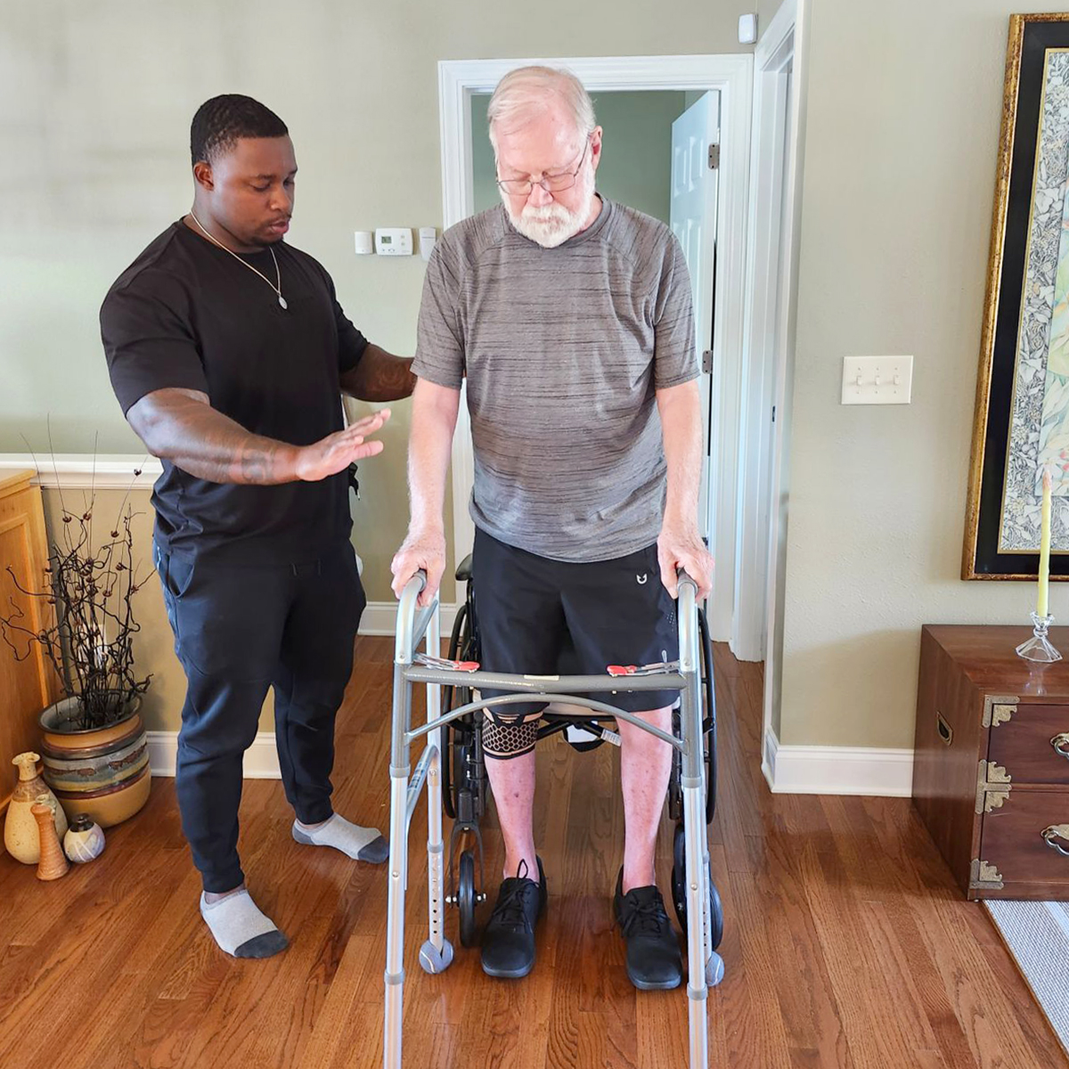 An older man uses a walker to stand up from a wheelchair while a younger man stands nearby, offering assistance in a home setting.