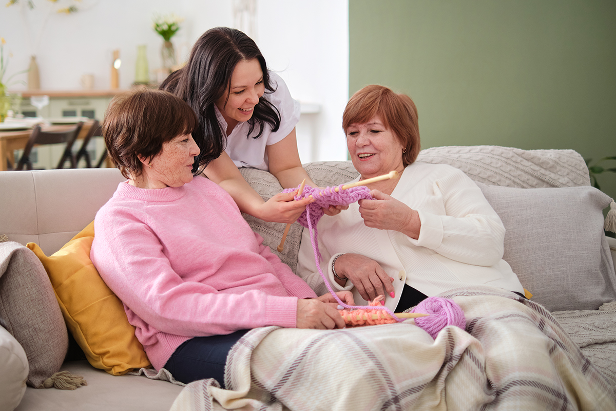 Two older women sit on a couch under a blanket, knitting with pink yarn, while a younger woman stands behind them, smiling and offering assistance.