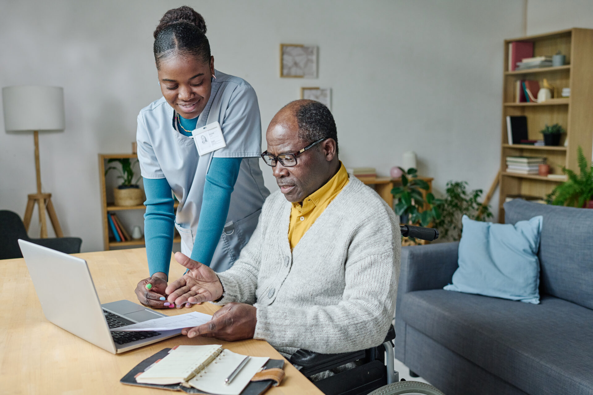 A caregiver stands next to an older man in a wheelchair as they look at paperwork and a laptop on a table in a living room.
