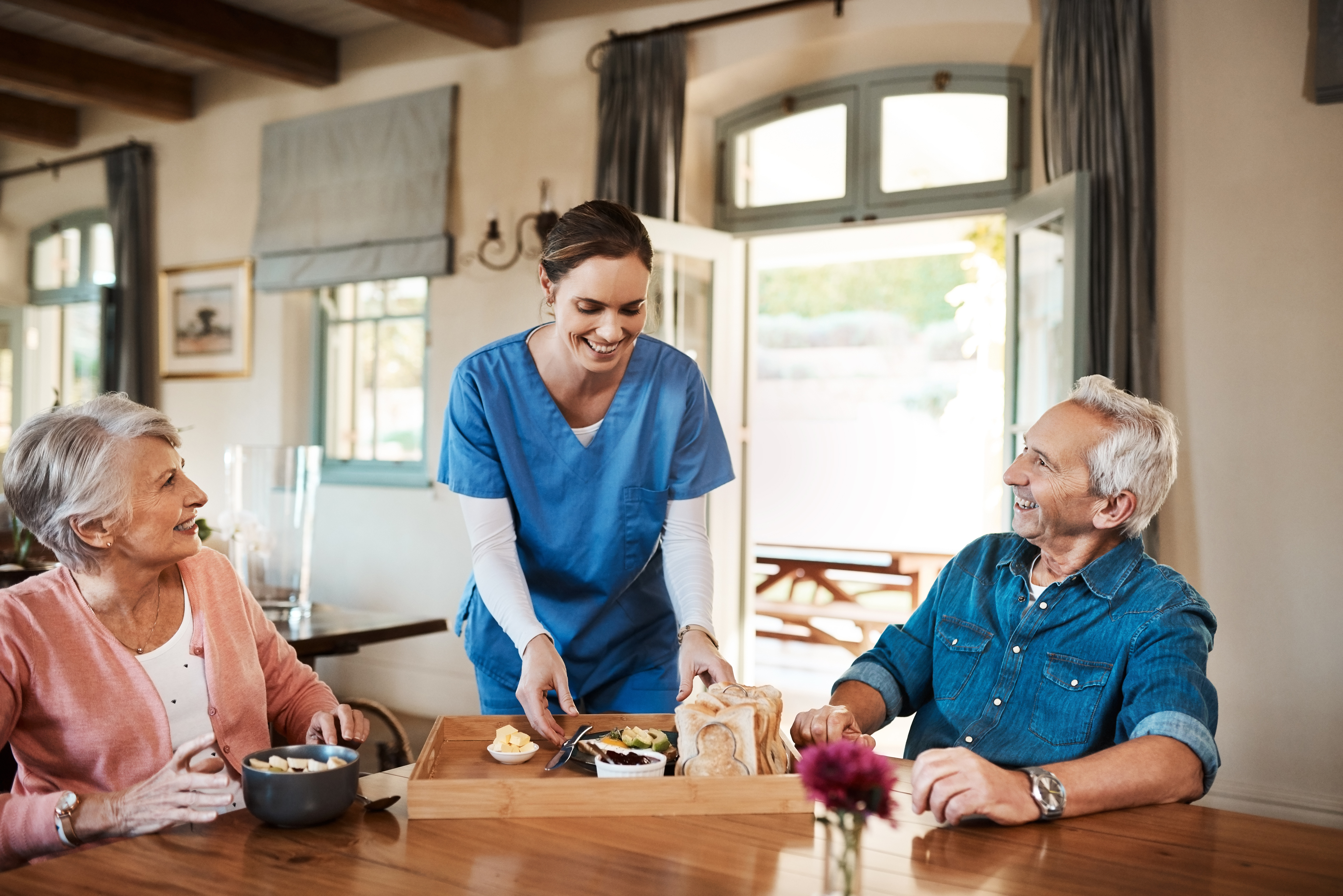 A caregiver in scrubs serves food to a smiling elderly couple sitting at a table in a bright, homey dining room.