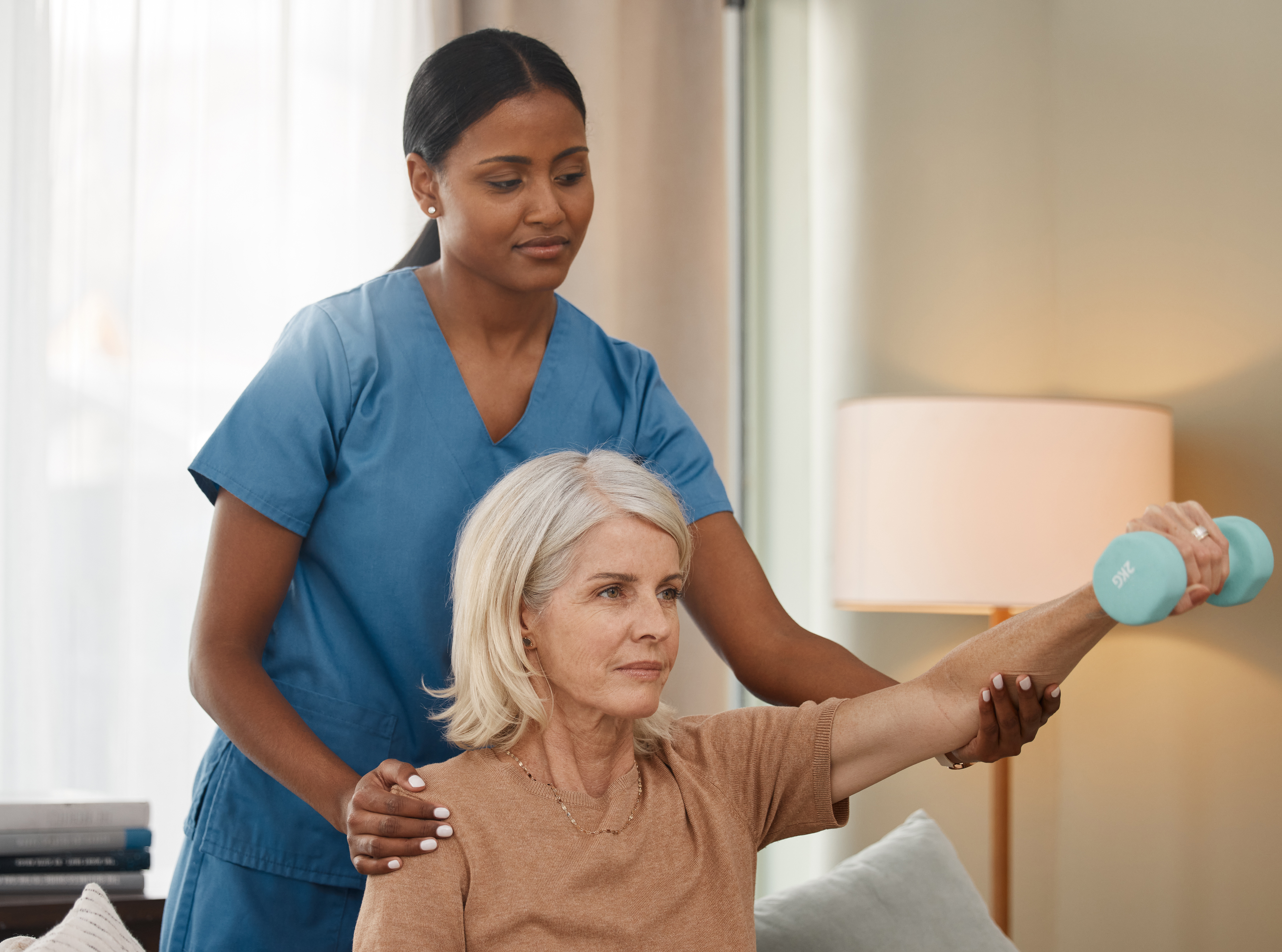 A healthcare worker assists an older woman with a physical therapy exercise using a dumbbell in a home setting.