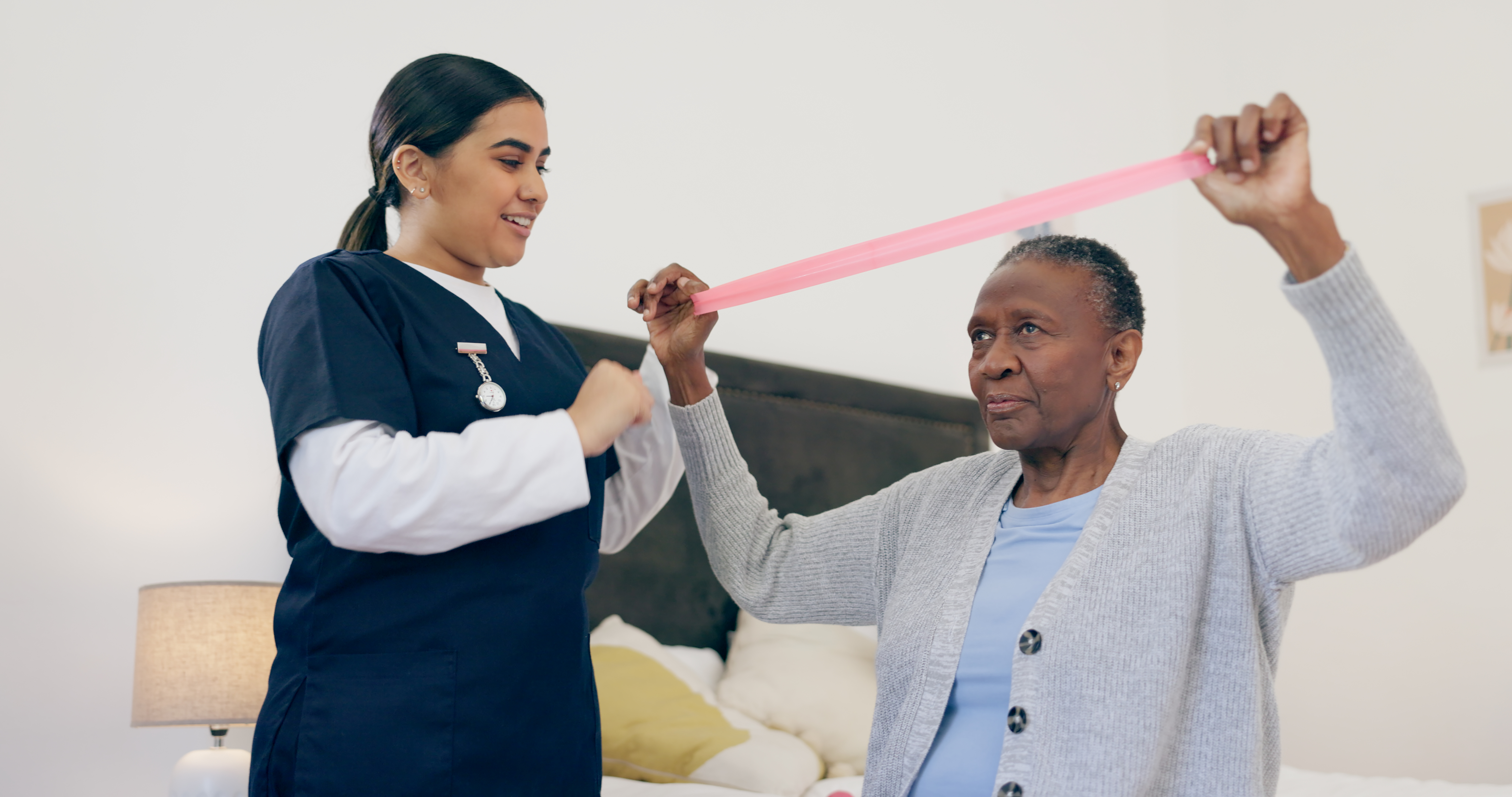 A nurse assists an older woman with arm exercises using a pink resistance band in a bedroom setting.